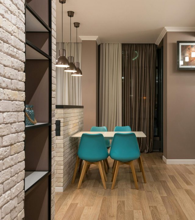 Turquoise chairs at table placed near window with curtains in modern spacious apartment with shelves and decorative white brick wall